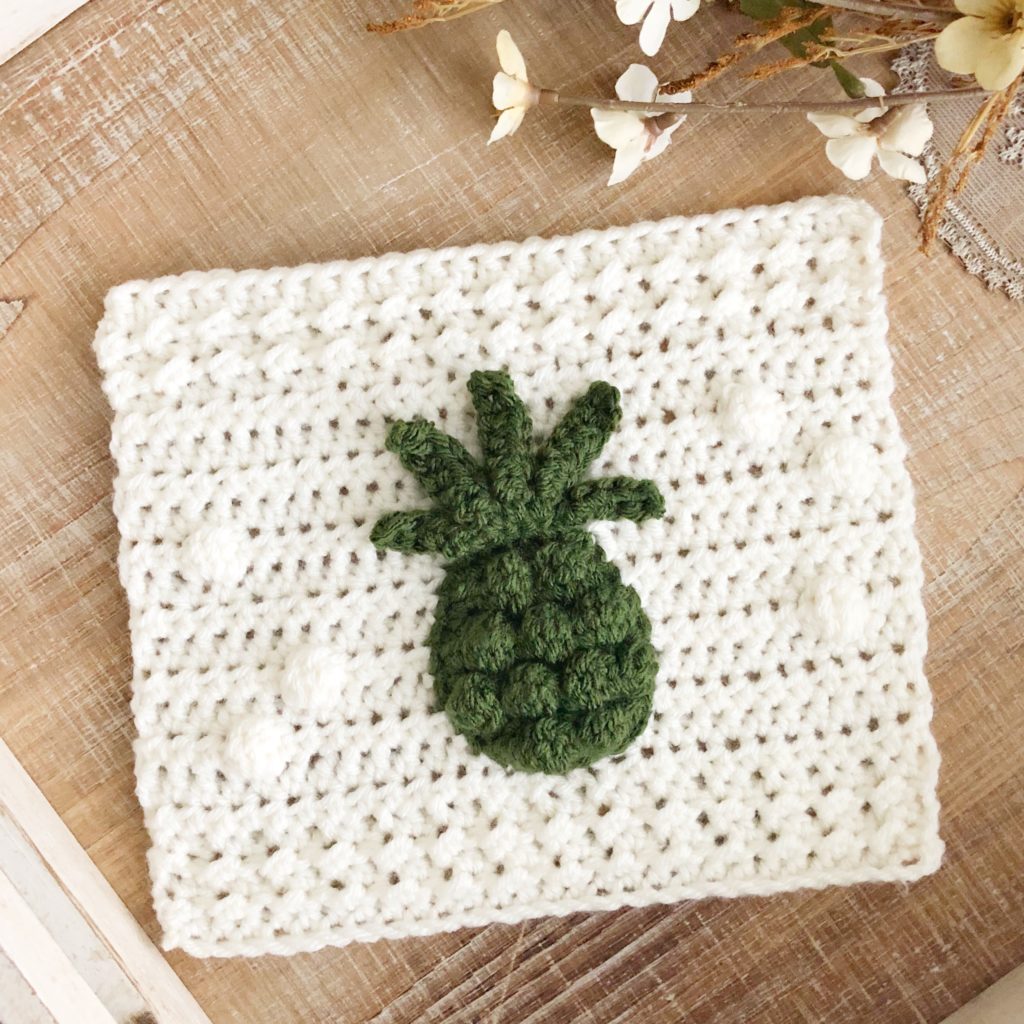 An off-white crochet square that is part of a blanket CAL with a dark green crochet pineapple applique sitting atop a neutral background accented by white flowers and coffee mug.
